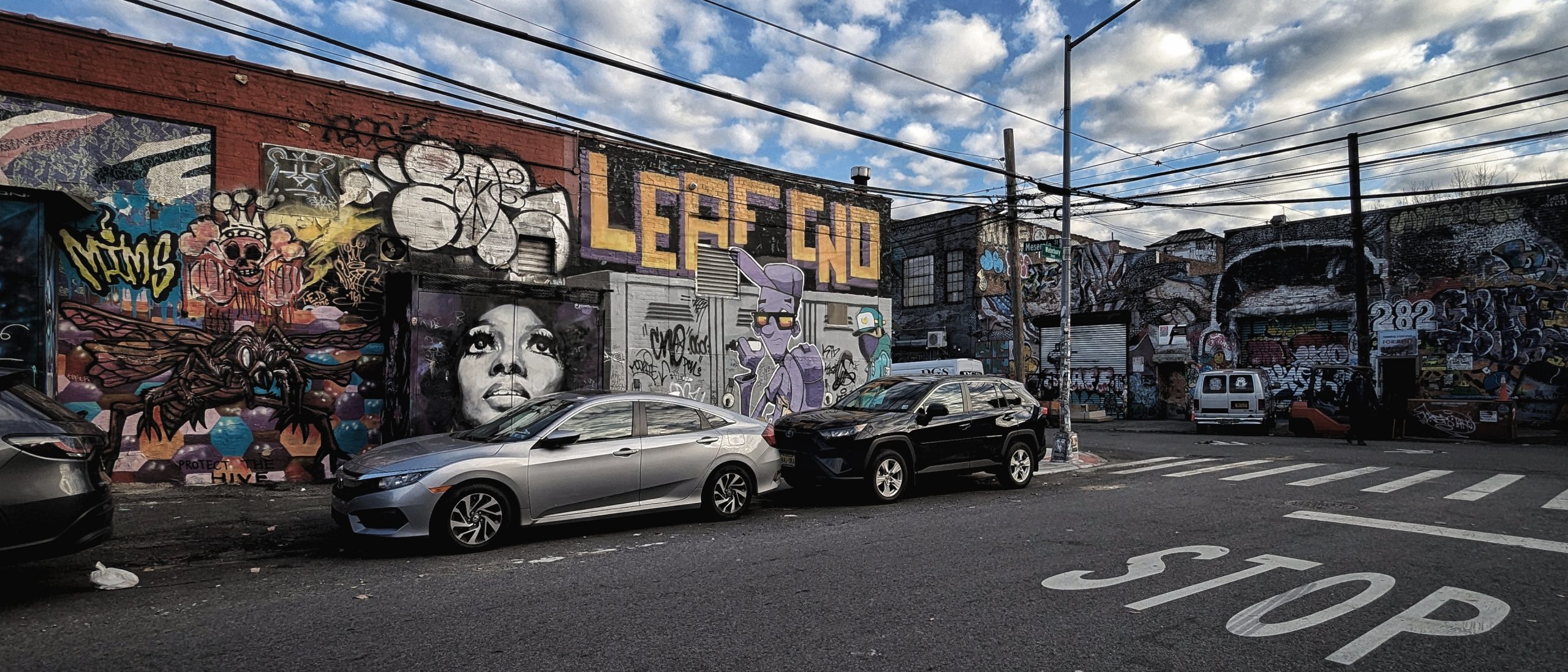 A wide-angle, atmospheric shot of a vibrant, graffiti-covered industrial street in Bushwick, Brooklyn. This image captures the raw, urban energy essential to our Private NYC Tours, illustrating the deep-focus, immersive experience of 'The Feature Presentation' as we explore the evolving textures of the city's outer boroughs.