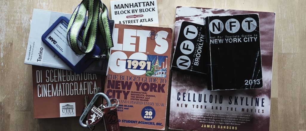 A flat-lay photograph on a wooden surface showing the curated tools of a NYC tour guide with a cinematic eye: a professional NYC tour guide license, a collection of vintage and modern city guidebooks, a film studies book, and a Torino football club keychain, representing the transition from a childhood in Italy to life in New York.