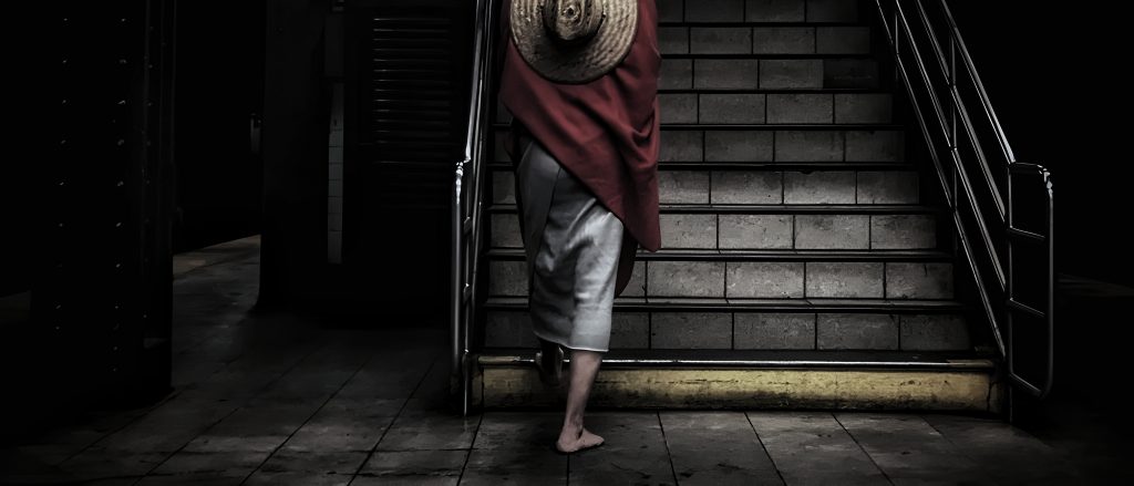 A cinematic, high-contrast photograph of a person walking up a dimly lit urban staircase, seen from behind. The subject is draped in a red fabric with a straw hat, creating a moody, mysterious atmosphere that mirrors the 'inutile' and narrative-driven nature of a GUIDA INUTILE NEW YORK tour.