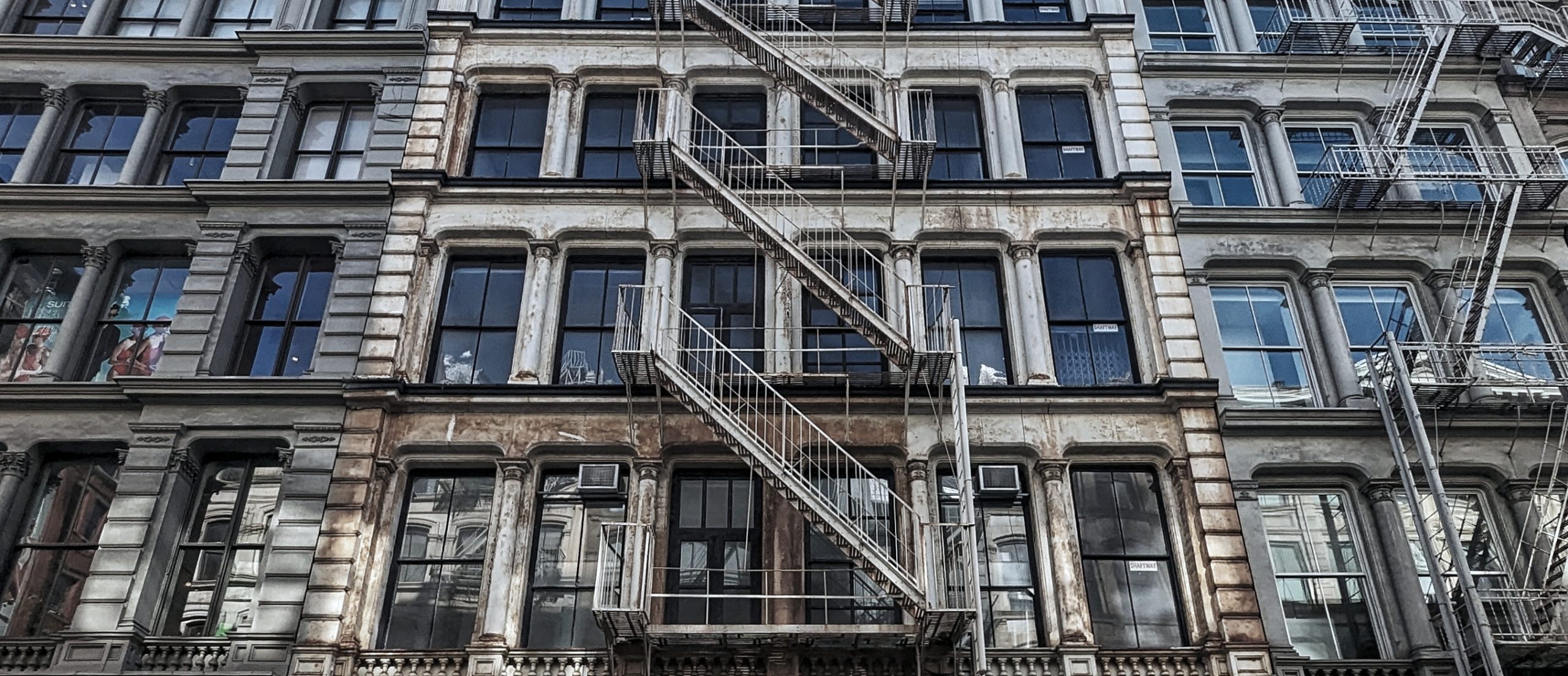 A cinematic, eye-level shot of a historic cast-iron building facade in New York City, featuring its distinctive rhythmic windows and intricate fire escapes. The image sets the scene for a focused, neighborhood-level exploration, capturing the iconic architectural textures that define New York’s 'B-side' character.
