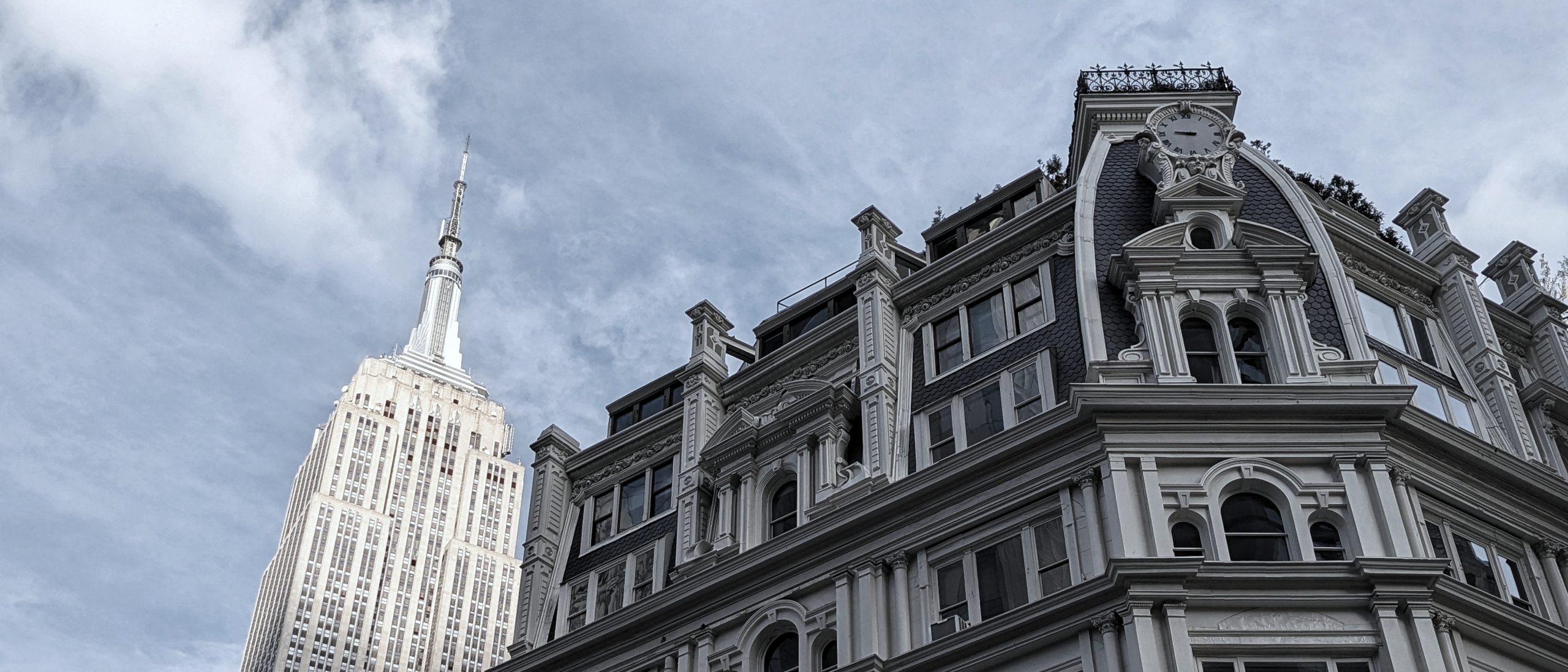 A cinematic, upward-looking shot contrasting the monumental peak of the Empire State Building with the ornate, historical architecture of a foreground building. This composition captures the essence of our Private NYC Tours—specifically 'The Short Subject'—by connecting the dots between iconic landmarks and the sophisticated, often overlooked architectural layers that exist right beside them.