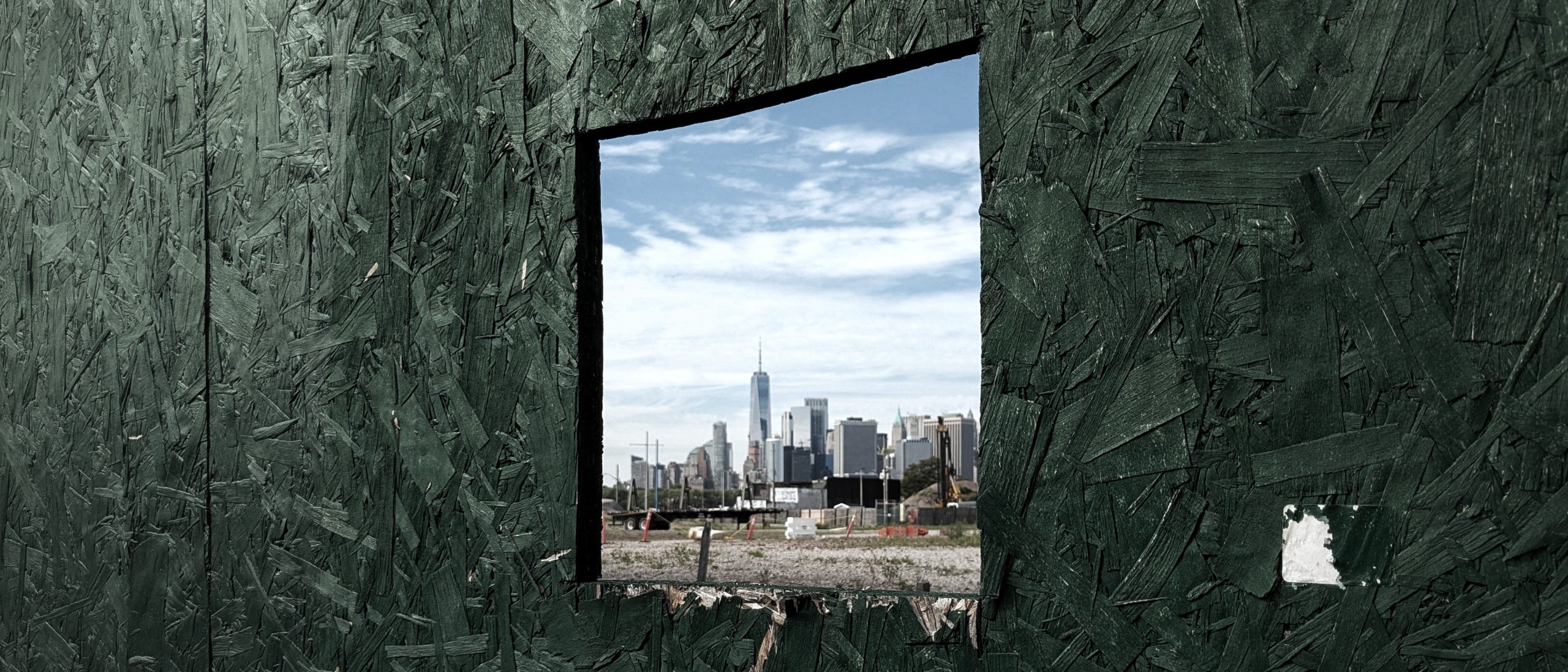 A cinematic frame of the Manhattan skyline viewed through a small, square cutout in a rough, green-painted wooden construction fence in Red Hook. This composition reflects the directorial approach of our Private NYC Tours, which prioritize the sophisticated 'Deep Cuts' and hidden architectural frames that reveal the true, living character of New York City.